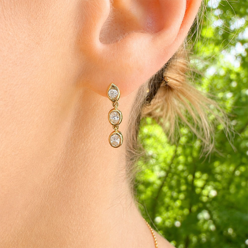 A close-up of a woman's ear wearing a 14K gold drop earring with three oval clear diamonds, set against a blurred green outdoor background.