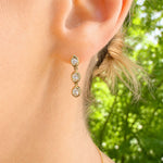 A close-up of a woman's ear wearing a 14K gold drop earring with three oval clear diamonds, set against a blurred green outdoor background.