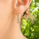 A close-up of a woman's ear wearing a 14K gold drop earring with three oval clear diamonds, set against a blurred green outdoor background.
