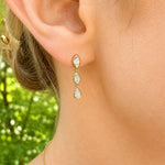 A close-up of a model's ear wearing a gold drop earring with three teardrop-shaped clear diamonds, set against a blurred green outdoor background.