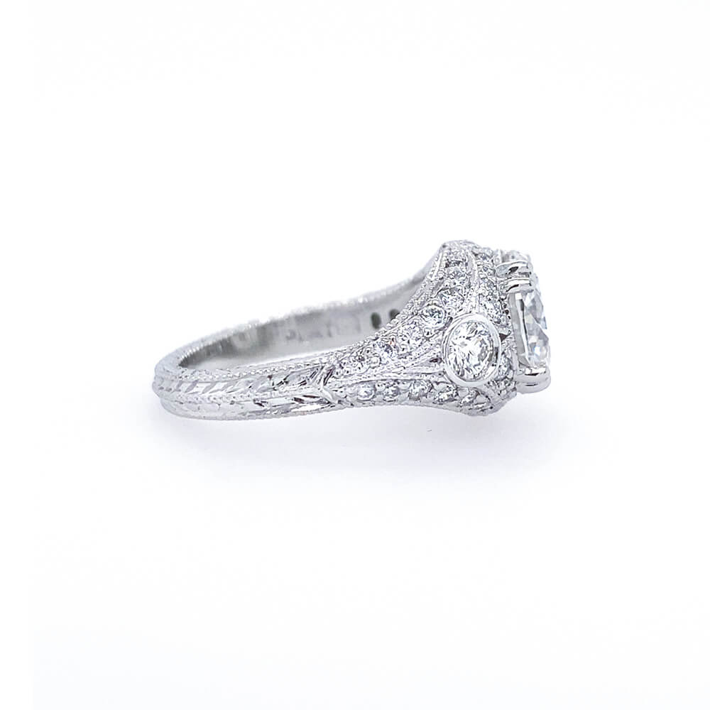 An antique-style diamond ring in platinum metal, featuring a central round diamond surrounded by smaller diamonds and a decorative engraved band, set against a plain white background.