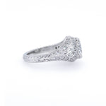 An antique-style diamond ring in platinum metal, featuring a central round diamond surrounded by smaller diamonds and a decorative engraved band, set against a plain white background.