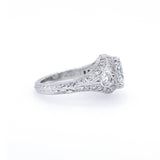 An antique-style diamond ring in platinum metal, featuring a central round diamond surrounded by smaller diamonds and a decorative engraved band, set against a plain white background.