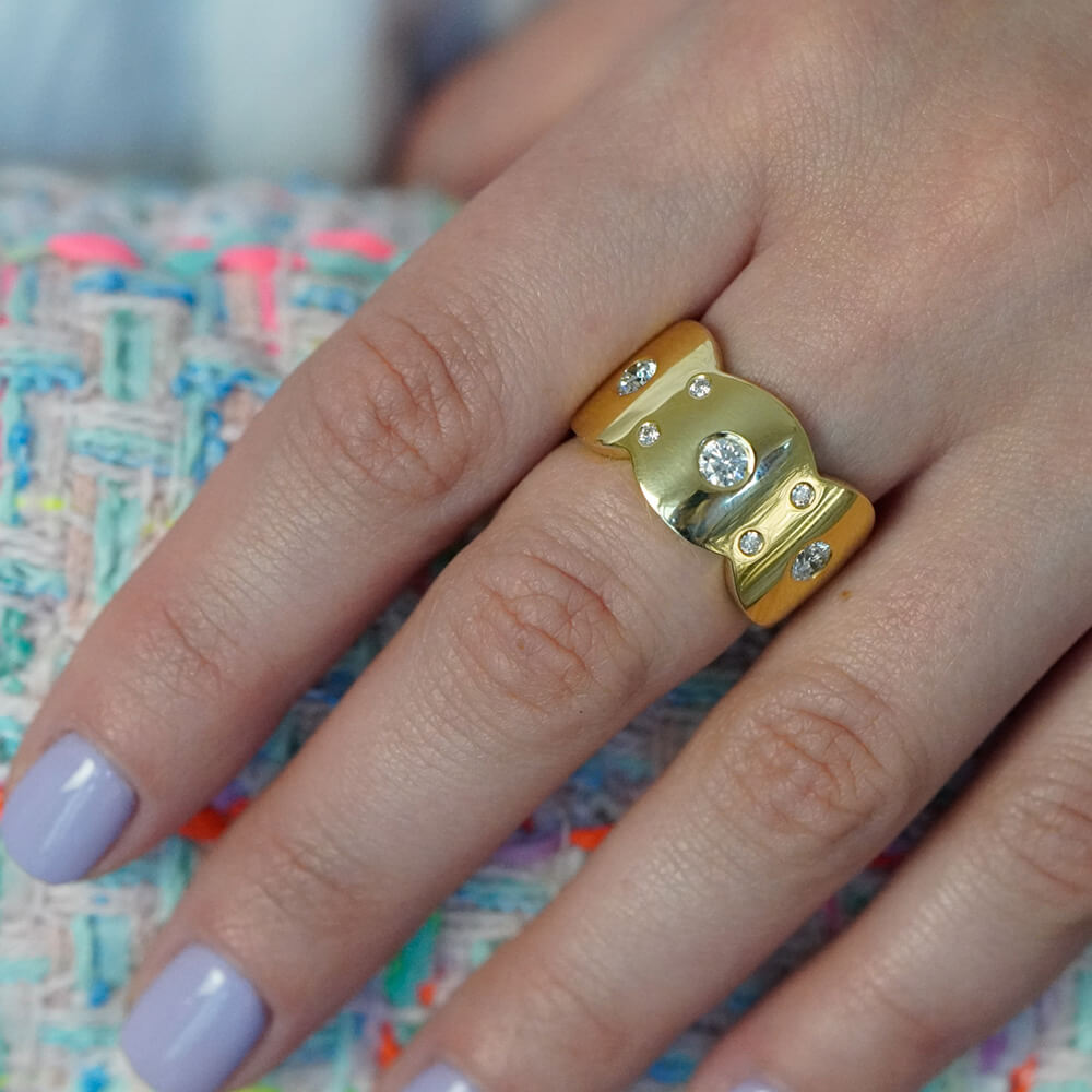 Gold scalloped edge ring with diamonds on a finger against a colorful fabric background