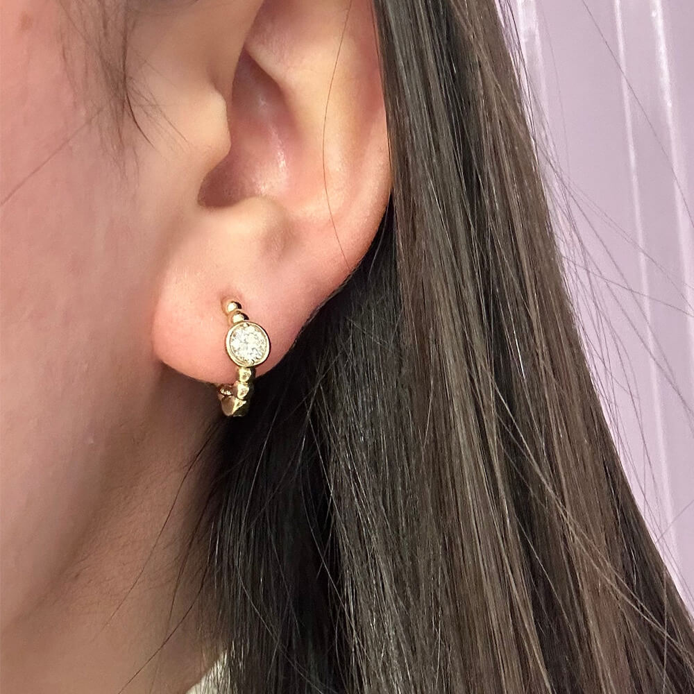 14K Gold earring with a small round bezel diamond worn by a woman with dark hair.