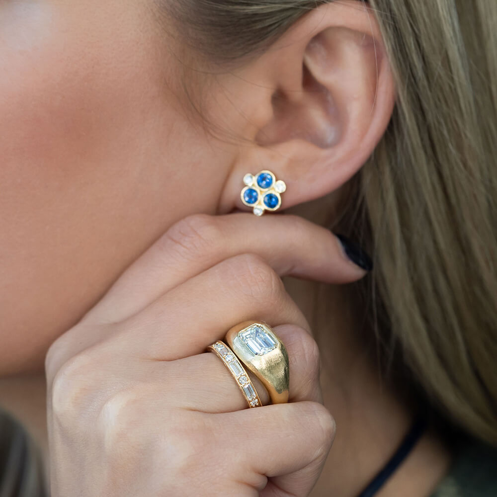 A close-up of a woman wearing an 18K yellow gold stud earring with three blue sapphires in a flower-like cluster, along with gold rings on her finger.
