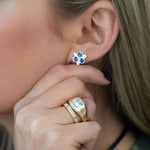 A close-up of a woman wearing an 18K yellow gold stud earring with three blue sapphires in a flower-like cluster, along with gold rings on her finger.