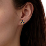 A close-up of a woman's ear wearing an 18K yellow gold stud earring with three round blue sapphires and smaller clear accent diamonds.