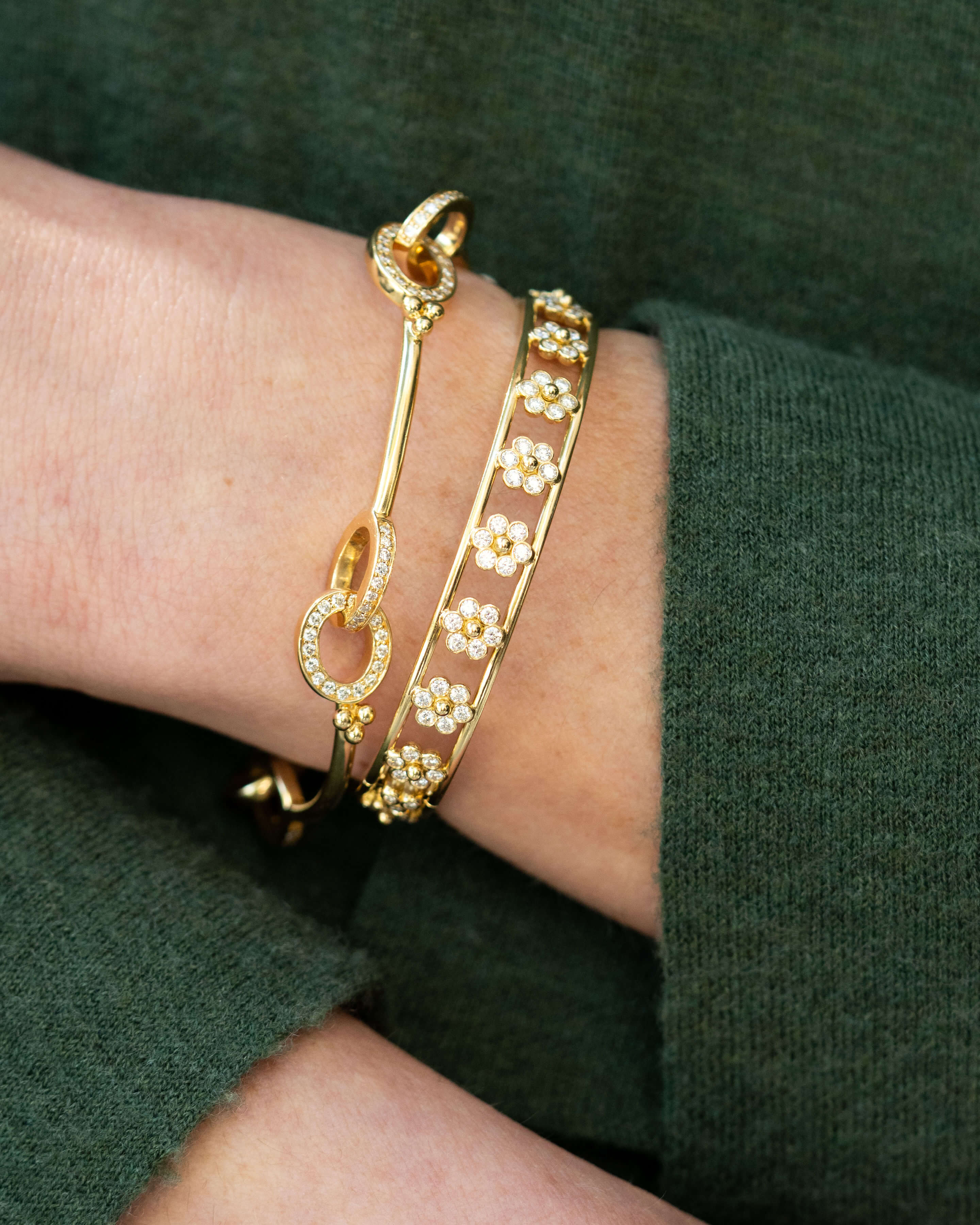 A close-up of a person's wrist wearing two gold bracelets: one with a heart-shaped clasp and one with a row of small flower motifs, set against a dark green knit top.