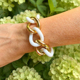 A close-up of a woman's wrist wearing a chunky chain-link bracelet made of alternating glossy white agate and polished 18K gold links, set against a background of green hydrangea flowers.