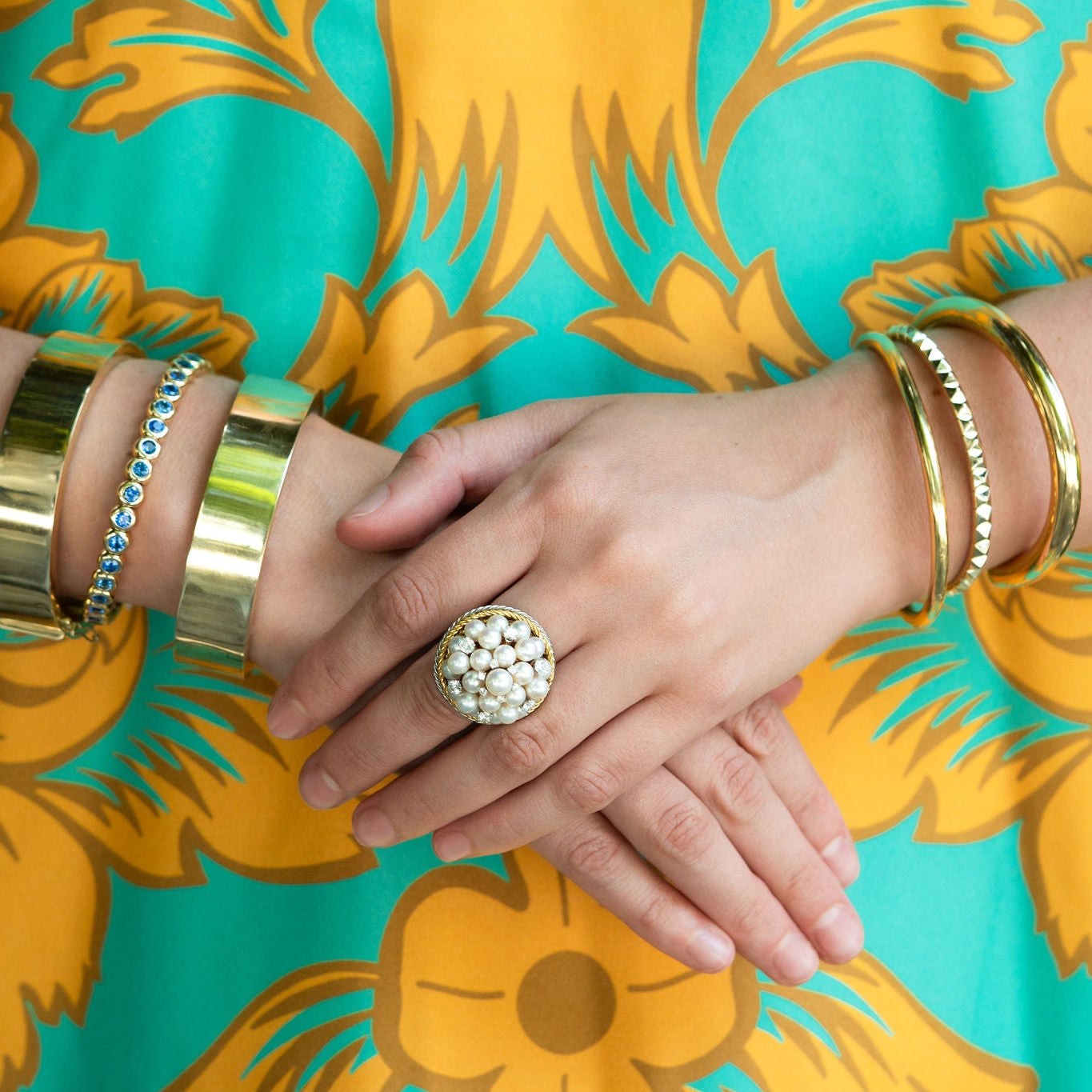 Close-up of woman's hands with gold bracelets and an estate Pearl ring against a floral-patterned fabric background.