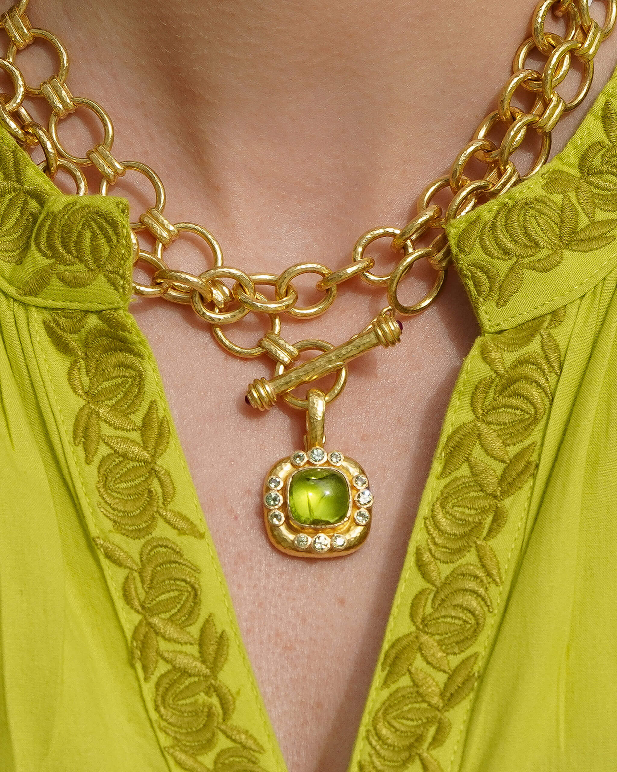 A close-up of a gold chain necklace with a toggle clasp and a square pendant featuring a green gemstone surrounded by small clear stones, worn by a model in a bright yellow-green embroidered top.