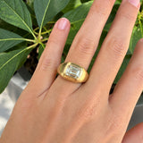 A gold signet ring with a rectangular emerald-cut clear diamond, worn on a woman's hand with green leaves in the background.