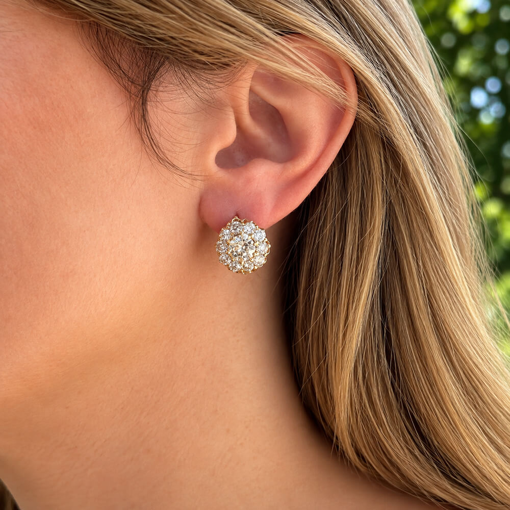 A close-up of a woman's ear wearing a stud earring with a cluster of clear diamonds in a rose gold setting, against a softly blurred outdoor background.