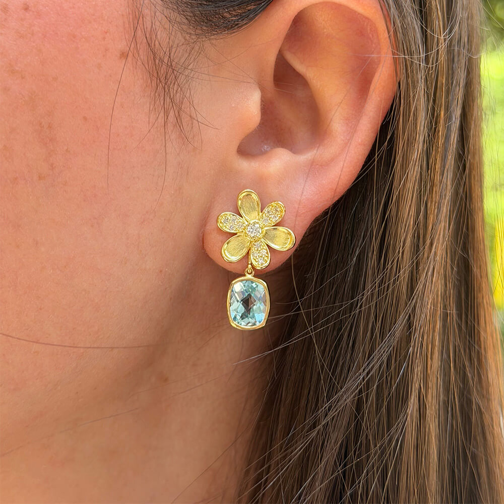 A close-up of a woman's ear wearing an 18K yellow gold flower-shaped stud earring with a dangling light blue topaz gemstone earring jacket