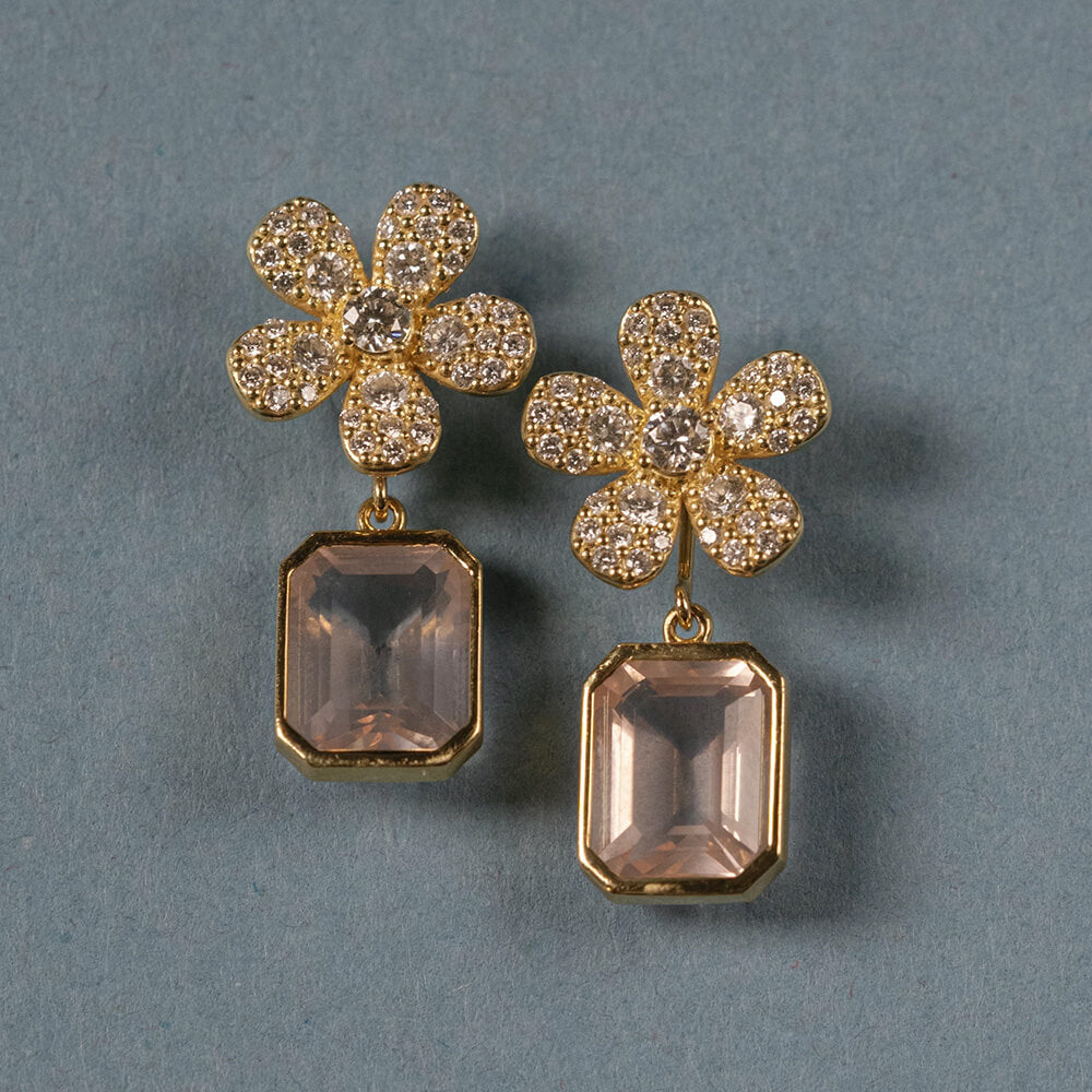 A pair of 18K yellow gold flower-shaped drop earrings with pavé-set clear diamonds and rectangular blush pink amethyst gemstone drops, displayed on a textured blue-gray background.