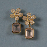 A pair of 18K yellow gold flower-shaped drop earrings with pavé-set clear diamonds and rectangular blush pink amethyst gemstone drops, displayed on a textured blue-gray background.