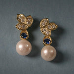 A pair of 18K yellow gold leaf-shaped drop earrings with clear pavé diamonds, a small round blue sapphire, and a large white pearl dangling below, set against a dark slate gray background.