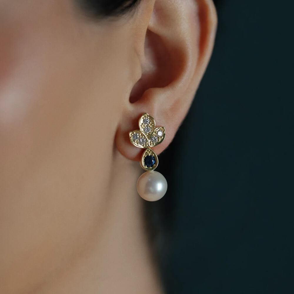 A close-up of a woman's ear wearing a gold leaf-shaped stud earring with clear diamonds, a dark blue teardrop sapphire gemstone, and a dangling white pearl.