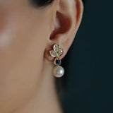 A close-up of a woman's ear wearing a gold leaf-shaped stud earring with clear diamonds, a dark blue teardrop sapphire gemstone, and a dangling white pearl.