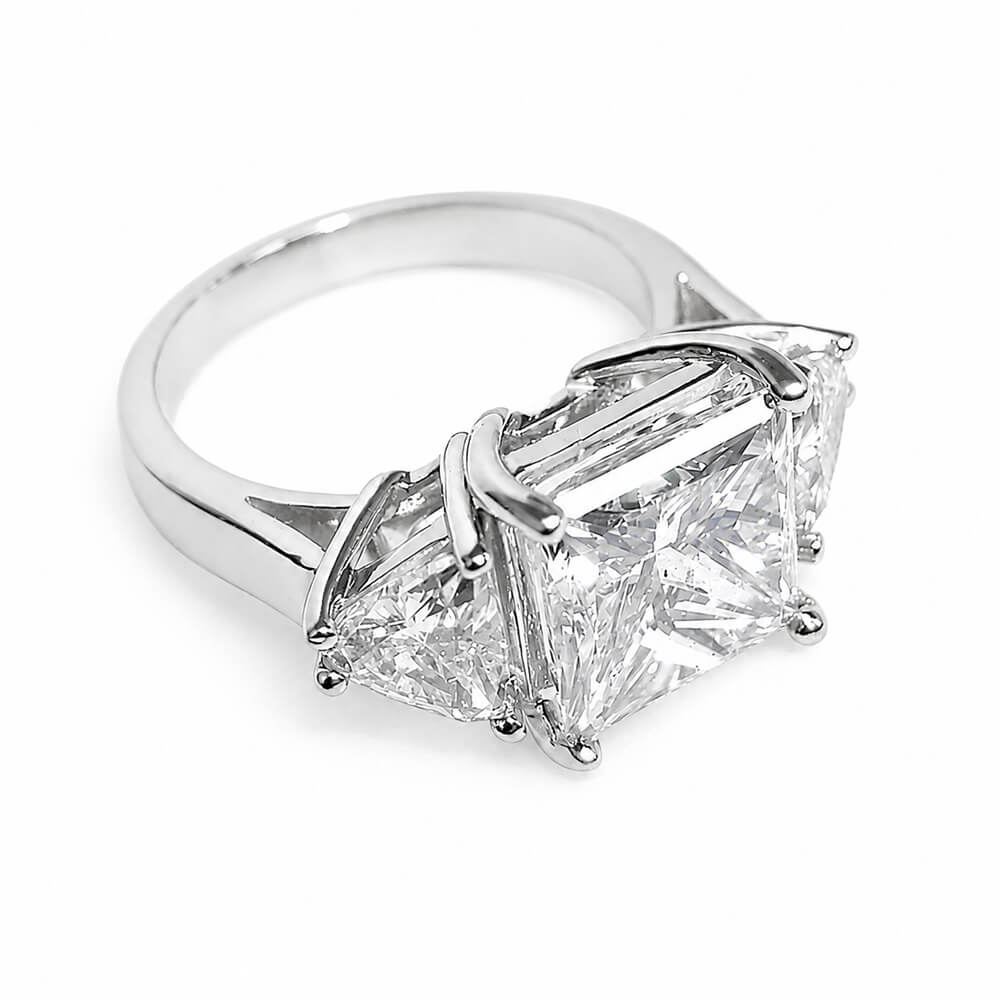 A three-stone diamond ring featuring a central princess-cut diamond flanked by two smaller trillion-cut diamonds in a platinum setting, shown on a plain white background.