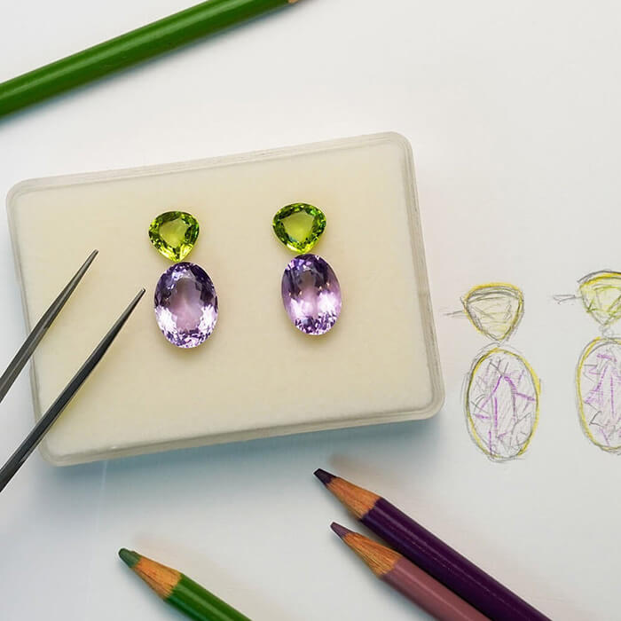Pair of custom designed earrings with purple and green precious gemstones on a white surface with jewelry design tools.