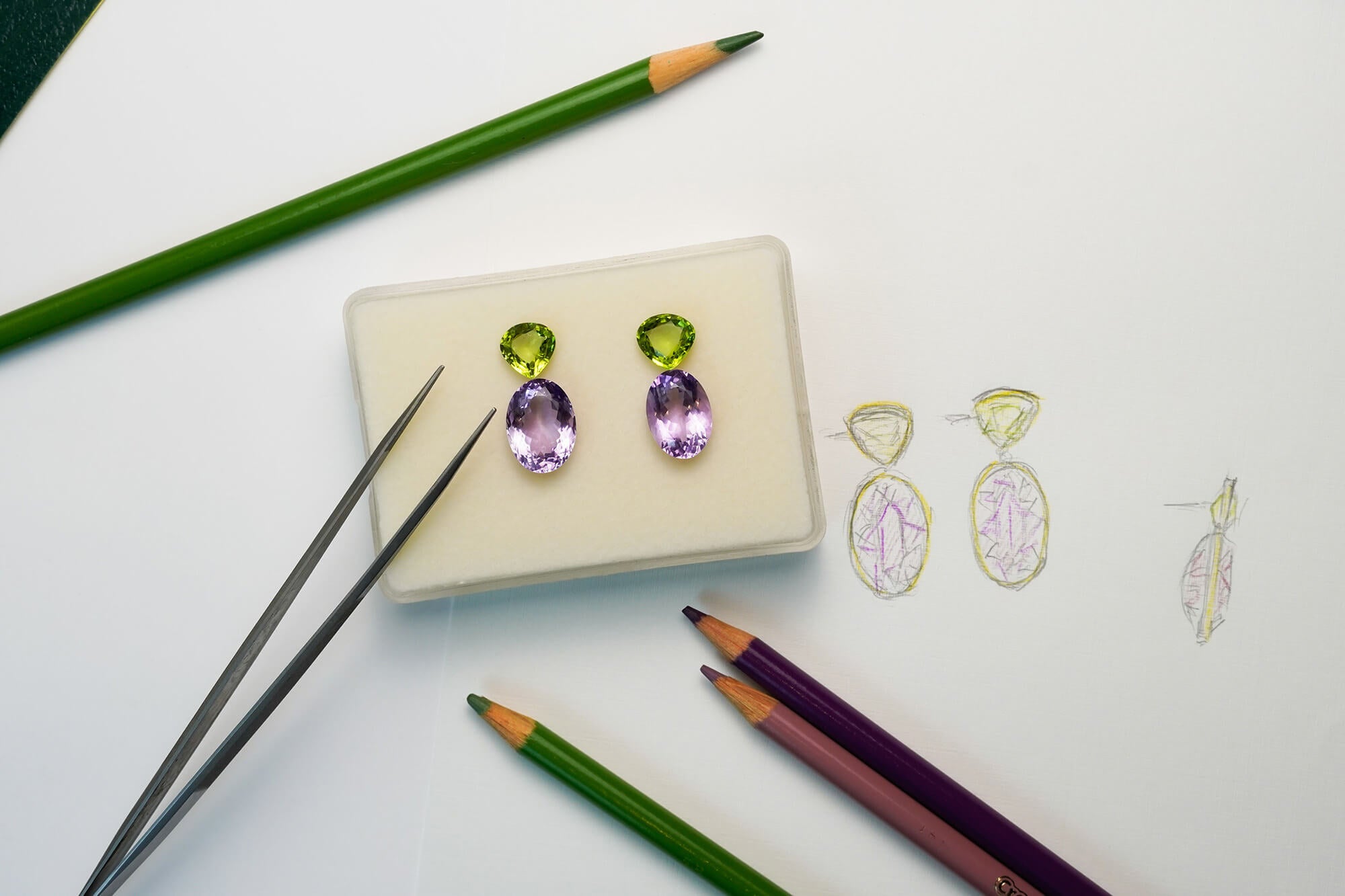 Custom earrings with green and purple gemstones with drawing tools nearby.