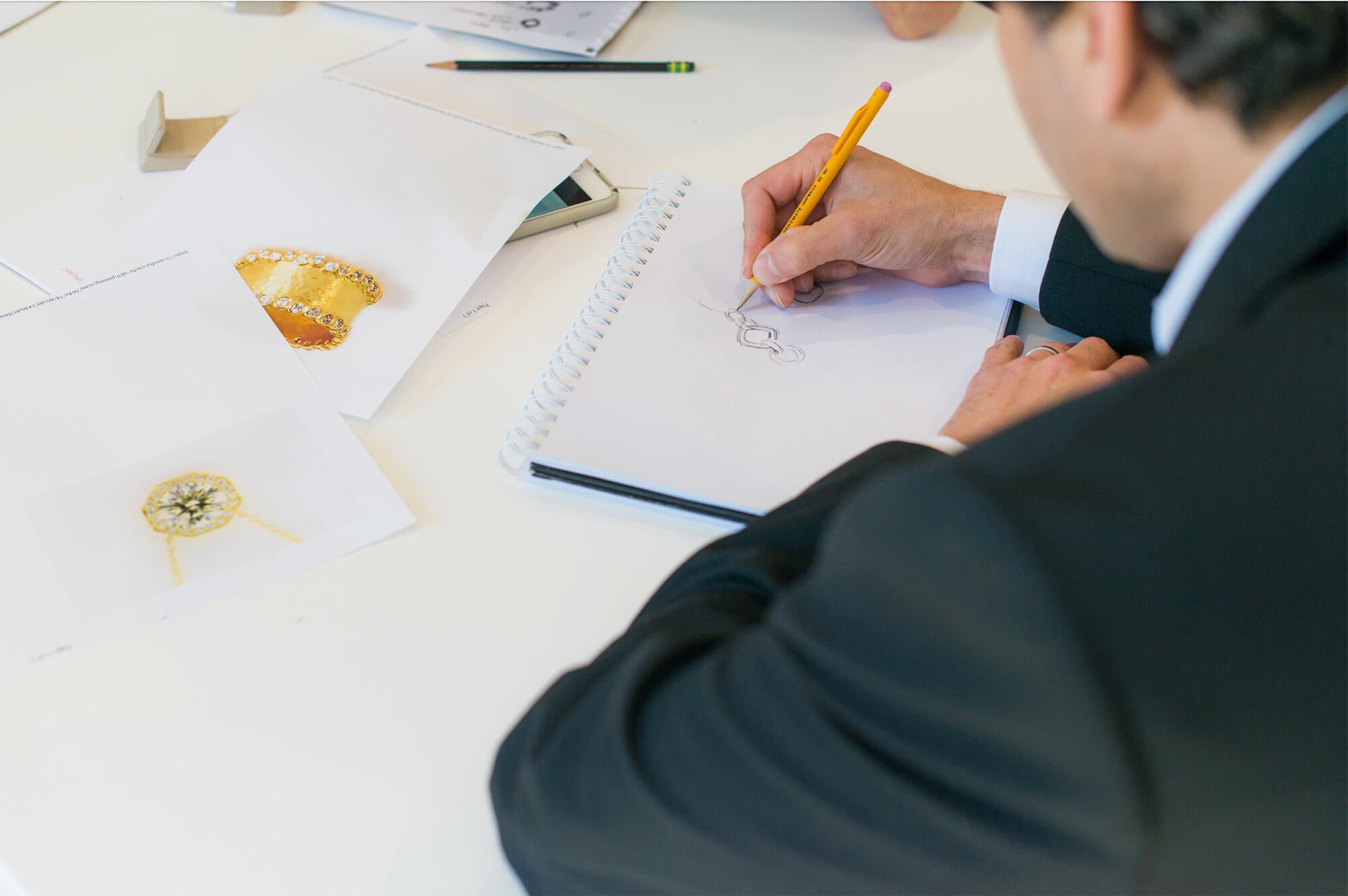 Jeweler drawing designs, surrounded by jewelry design sketches on a table.
