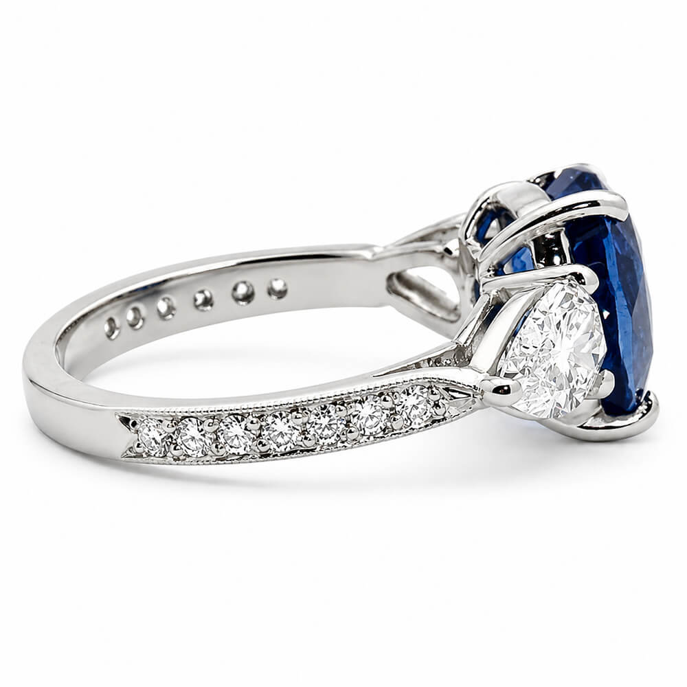 A platinum ring featuring a cushion cut blue sapphire and two pear shaped diamonds, with a pavé-set diamond band, shown on a white background.
