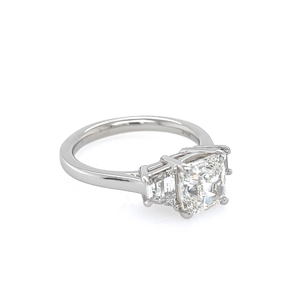 A ring featuring a large cushion-cut diamond center stone with two smaller trapezoid diamonds on either side, set in a polished platinum band against a plain white background.