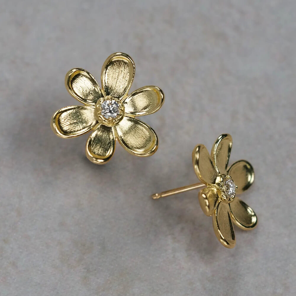 A pair of 18K yellow gold flower stud earrings with a small clear diamond in the center, displayed on a textured light gray surface.
