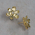 A pair of 18K yellow gold flower stud earrings with a small clear diamond in the center, displayed on a textured light gray surface.