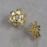 A pair of 18K yellow gold flower stud earrings with a small clear diamond in the center, displayed on a textured light gray surface.