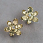 A pair of 18K yellow gold flower-shaped stud earrings with a small clear round diamond in the center, displayed on a neutral gray background.