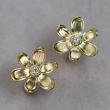 A pair of 18K yellow gold flower-shaped stud earrings with a small clear round diamond in the center, displayed on a neutral gray background.