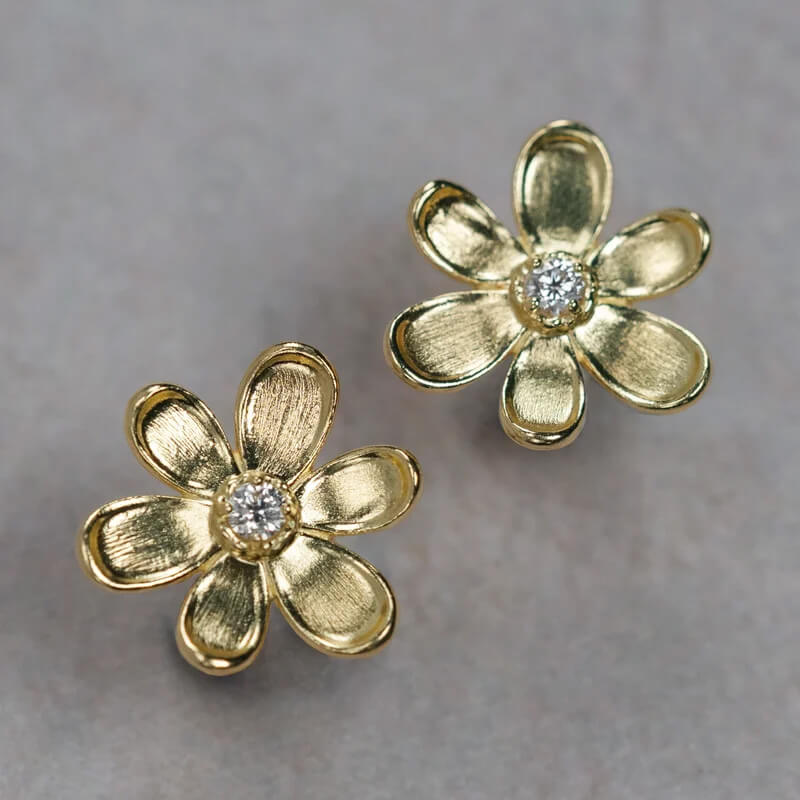 A pair of 18K yellow gold flower-shaped stud earrings with a small clear round diamond in the center, displayed on a neutral gray background.