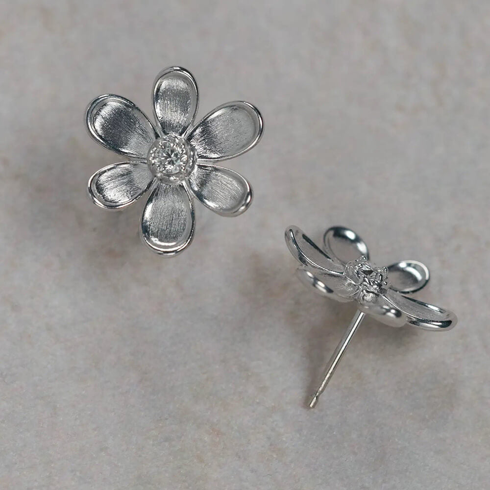 A pair of 18K white gold flower stud earrings, with one earring shown from the front displaying a textured five-petal design and a clear center diamond, and the other shown from the side/back profile. They are displayed on a light beige stone surface.
