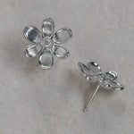 A pair of 18K white gold flower stud earrings, with one earring shown from the front displaying a textured five-petal design and a clear center diamond, and the other shown from the side/back profile. They are displayed on a light beige stone surface.