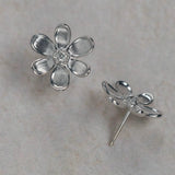 A pair of 18K white gold flower stud earrings, with one earring shown from the front displaying a textured five-petal design and a clear center diamond, and the other shown from the side/back profile. They are displayed on a light beige stone surface.