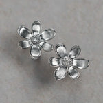 A pair of 18K white gold flower stud earrings with textured petals and a small clear diamond center, displayed on a neutral light gray surface.