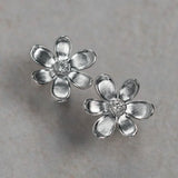 A pair of 18K white gold flower stud earrings with textured petals and a small clear diamond center, displayed on a neutral light gray surface.