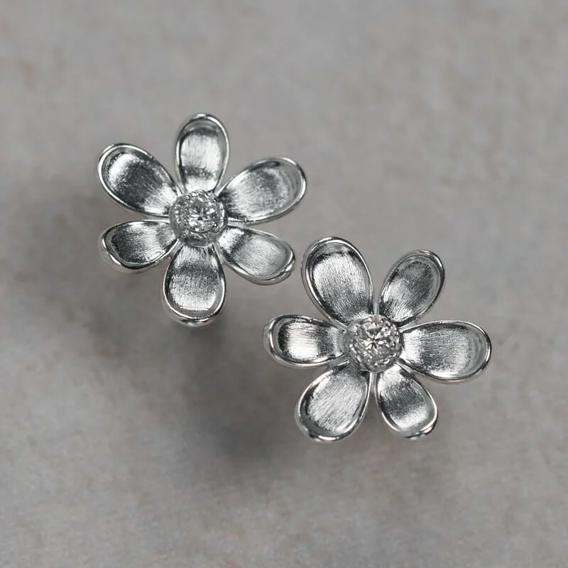 A pair of 18K white gold flower stud earrings with textured petals and a small clear diamond center, displayed on a neutral light gray surface.