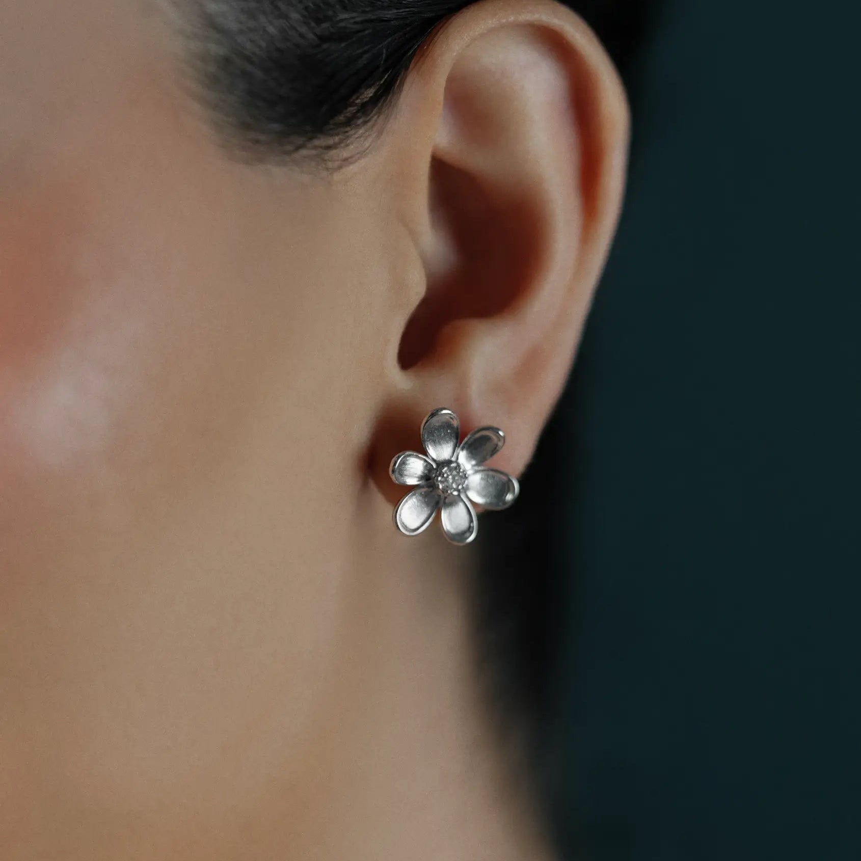 A close-up of a woman's ear wearing an 18K white gold flower-shaped diamond stud earring against a dark teal background.