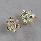 A pair of 18K yellow gold flower-shaped stud earrings with clear diamond accents in the center and along the petals, displayed on a light gray textured surface.