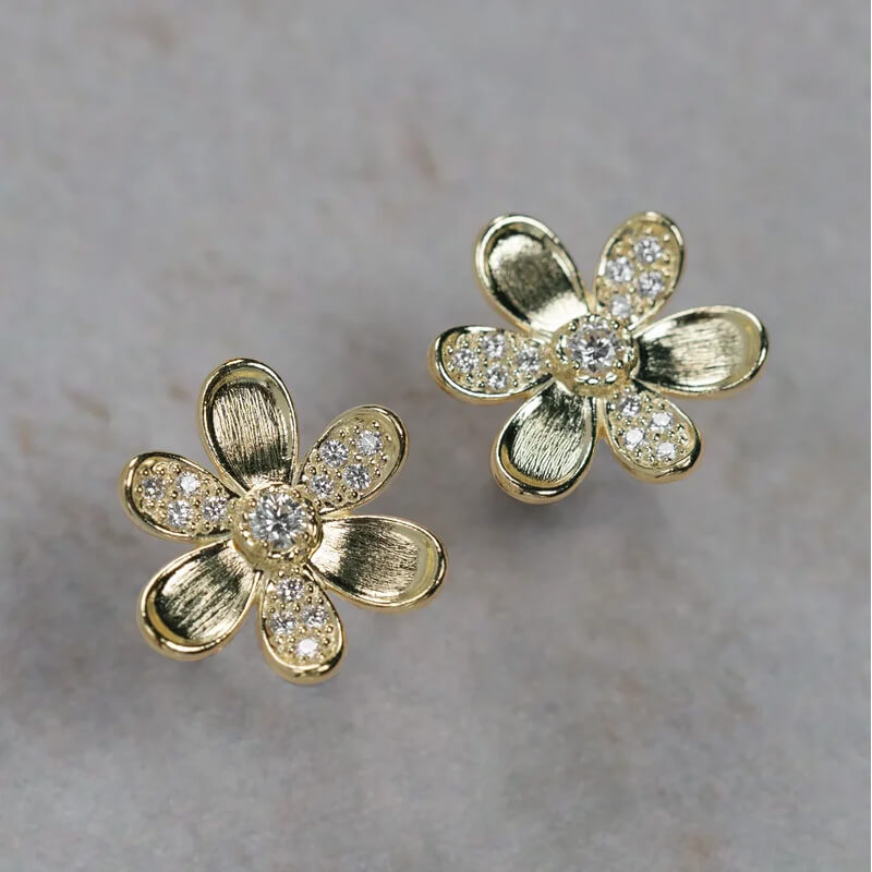 A pair of 18K yellow gold flower-shaped stud earrings with clear diamond accents in the center and along the petals, displayed on a light gray textured surface.
