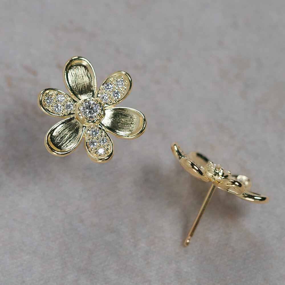 A pair of 18K yellow gold flower stud earrings with clear pavé-set stones, shown on a neutral textured gray background. One earring is shown from the front, displaying the textured gold petals and central round stone, while the other is shown from the side/back, displaying the post and backing.