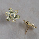 A pair of 18K yellow gold flower stud earrings with clear pavé-set stones, shown on a neutral textured gray background. One earring is shown from the front, displaying the textured gold petals and central round stone, while the other is shown from the side/back, displaying the post and backing.