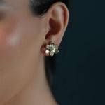 A close-up of a woman's ear wearing a small 18K yellow gold flower stud earring with clear diamonds, against a dark navy background.