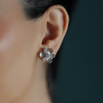 A close-up of a woman's ear wearing a small 18K white gold flower-shaped stud earring with textured petals and clear pave diamonds.