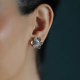 A close-up of a woman's ear wearing a small 18K white gold flower-shaped stud earring with textured petals and clear pave diamonds.